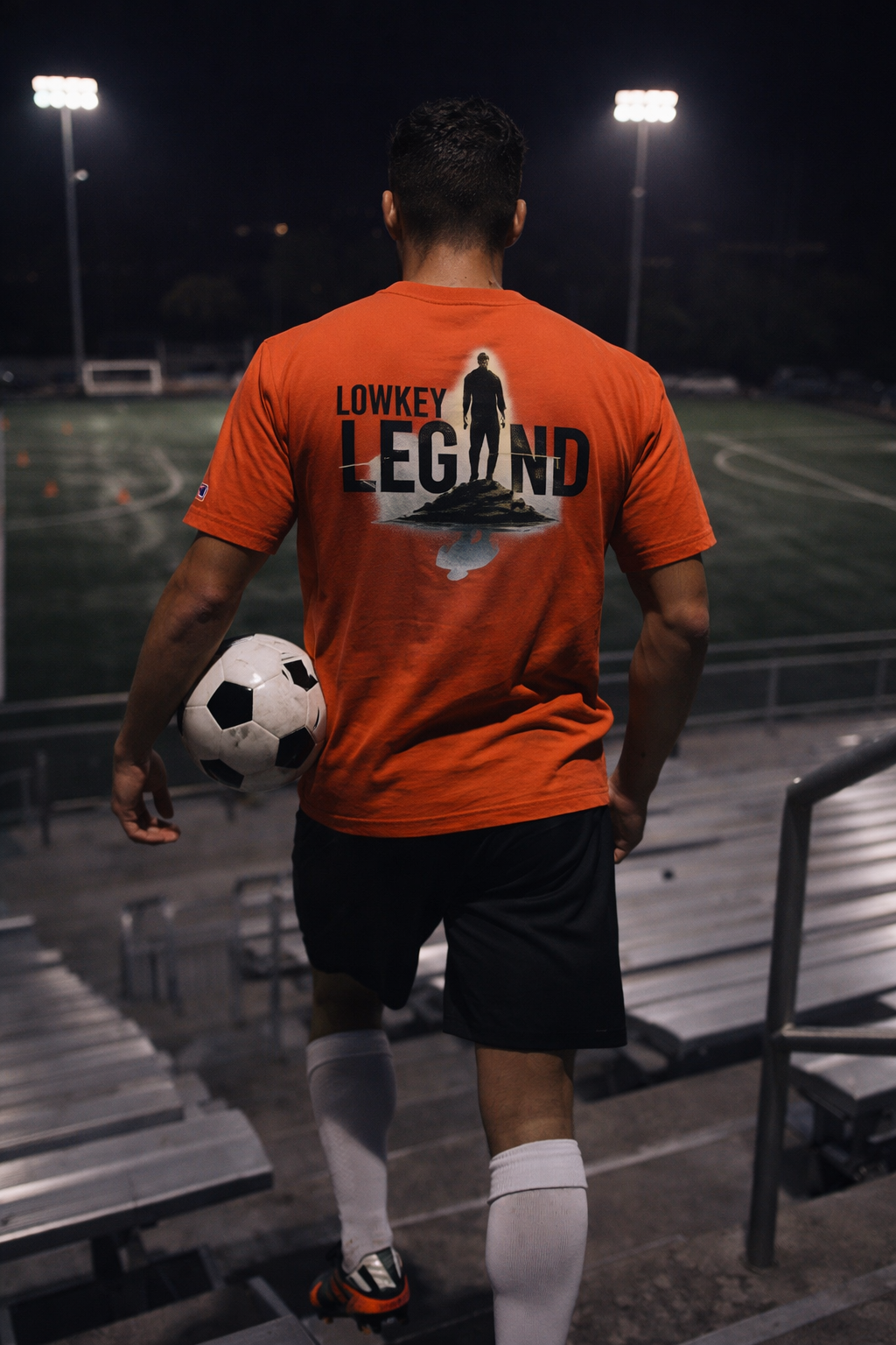 Person wearing an orange 'LOWKEY LEGEND' t-shirt holding a soccer ball on a sports field at night.